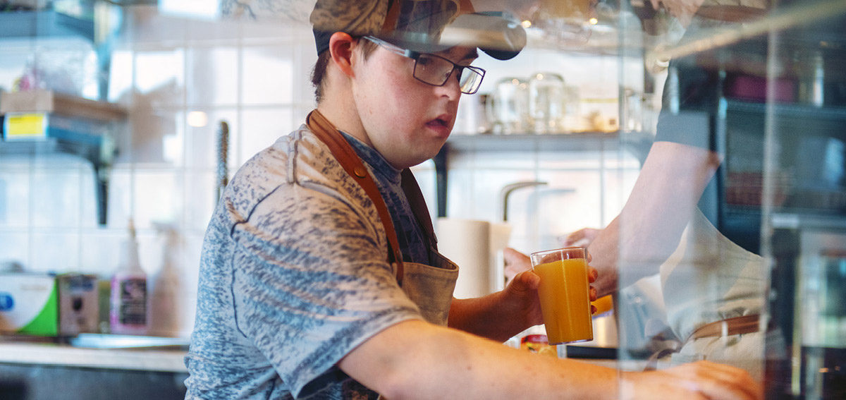 Young male adult living with down syndrome is working at a cafe and holding orange juice
