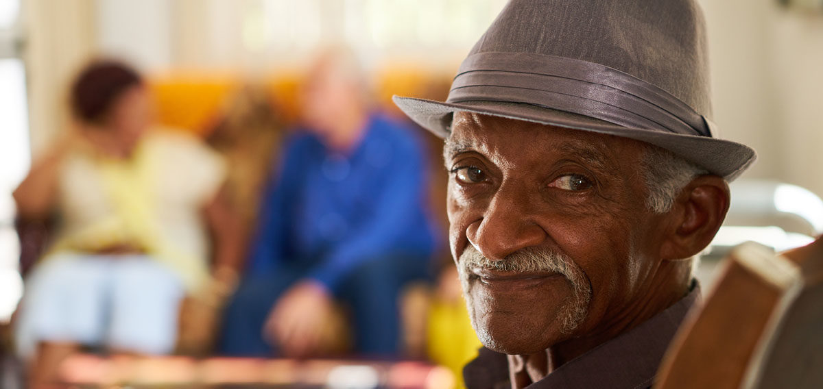 Older black man forcefully smiling at camera with hat on while he's indoors with two other people
