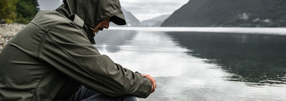 Man squatting next to river with a raincoat on, looking down contemplating