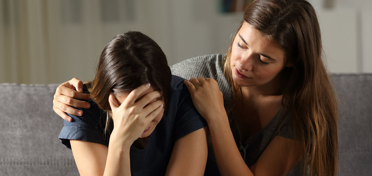 Young female consoling female friend whose crying with her hand covering her face on a couch indoors