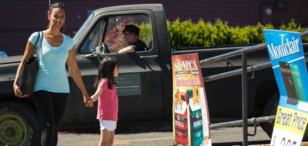 Young female child starting at tobacco ad outside of convenience store with mom while a truck drives past