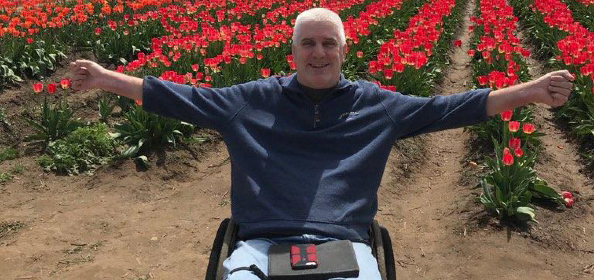 Older male in wheelchair sitting in flower field smiling with arms wide open towards the camera