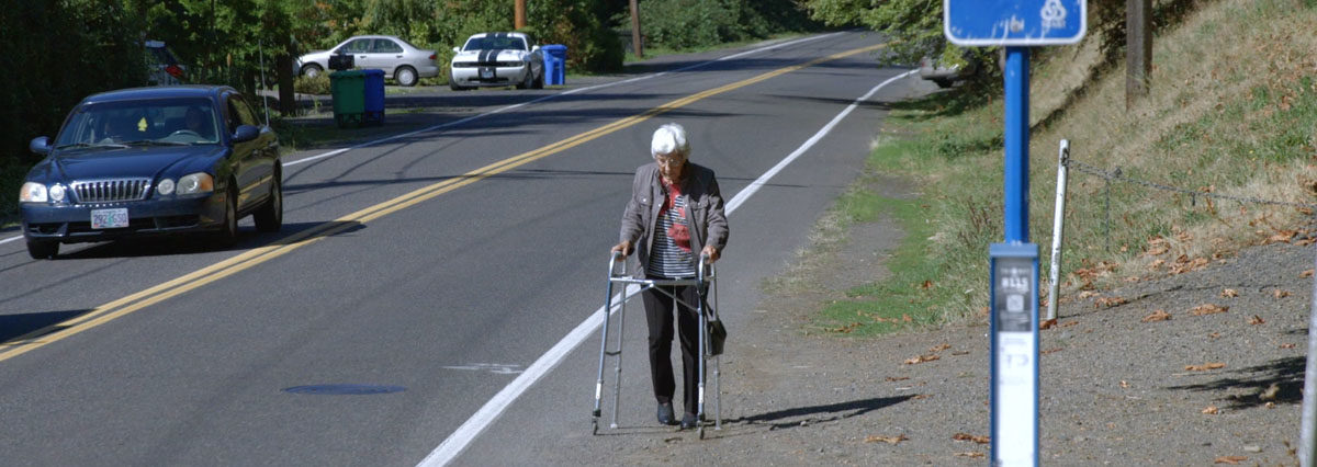 Older female adult walking on the side of a busy road towards a bus stop with a walker