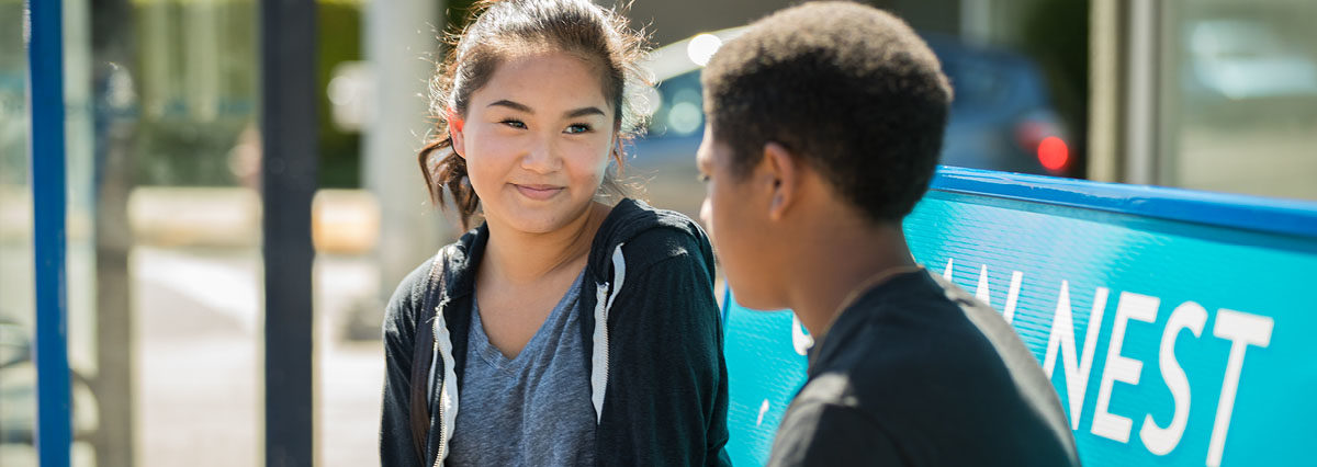 An Asian female teen smiling at a Black male teen while they sit outside at a bus stop
