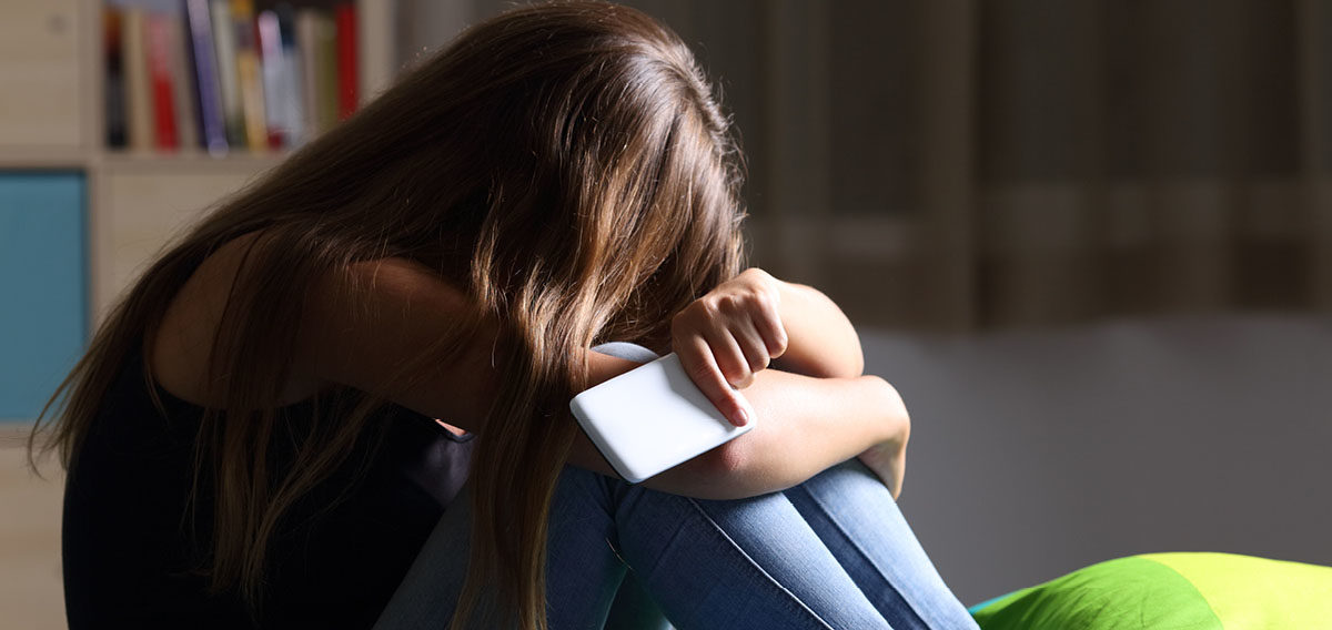 Female teen sitting in her room with her head in her hands and knees, covering her face holding her cellphone