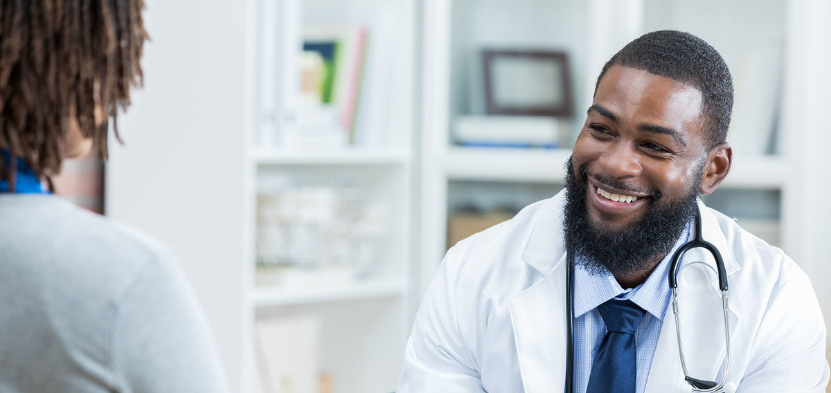 Black doctor with beard smiling at Black female patient inside doctor's office