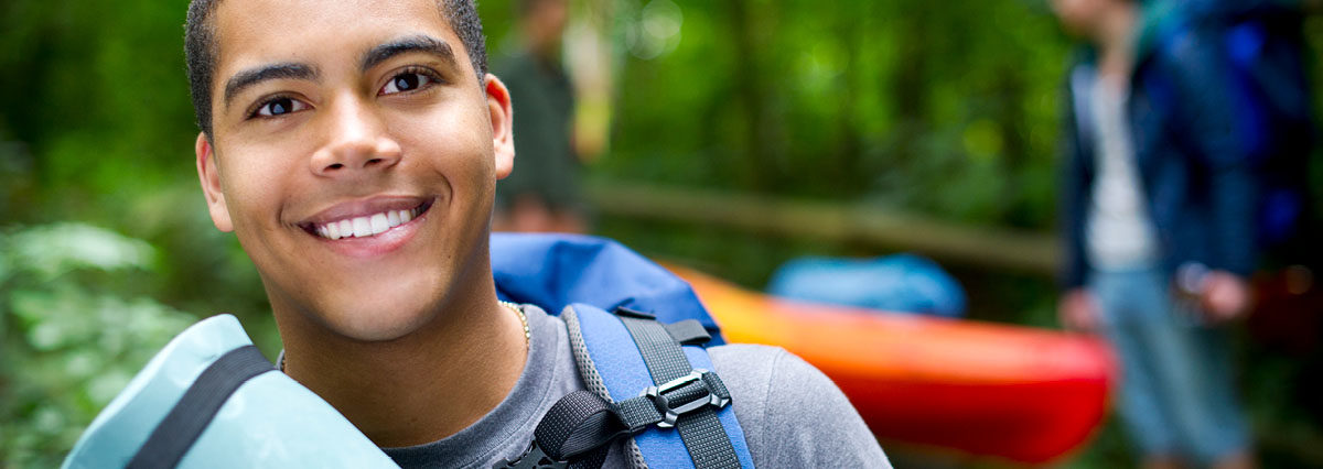 Black teen with camping goods smiling at camera while other teens in the background carry other camping items