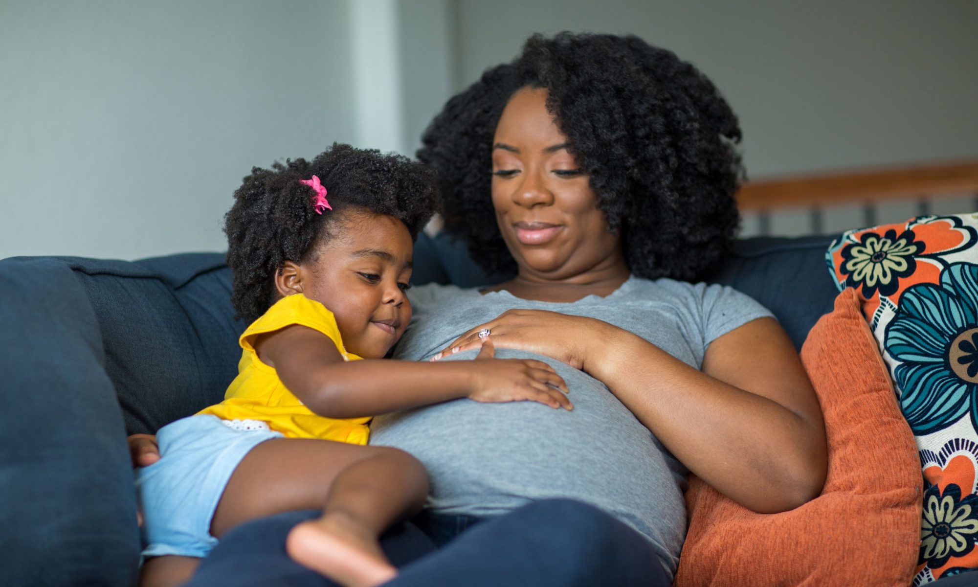 Black mother touching her pregnant stomach with her toddler daughter, sitting on couch inside