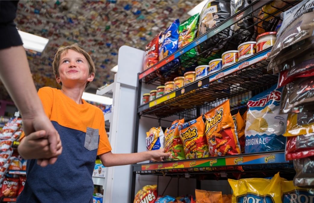 Young male kid grabbing Cheetos from large shelf of junk food products, smiling at mom while he holds her hand