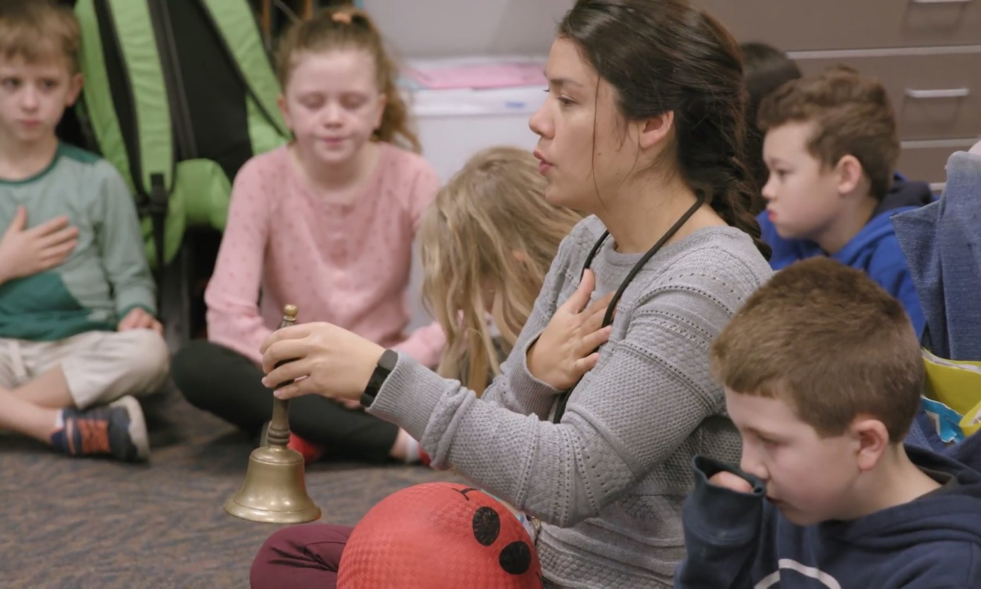 Teacher holding her chest and bell, teaching breathing exercises to students who are sitting on the floor in a classroom